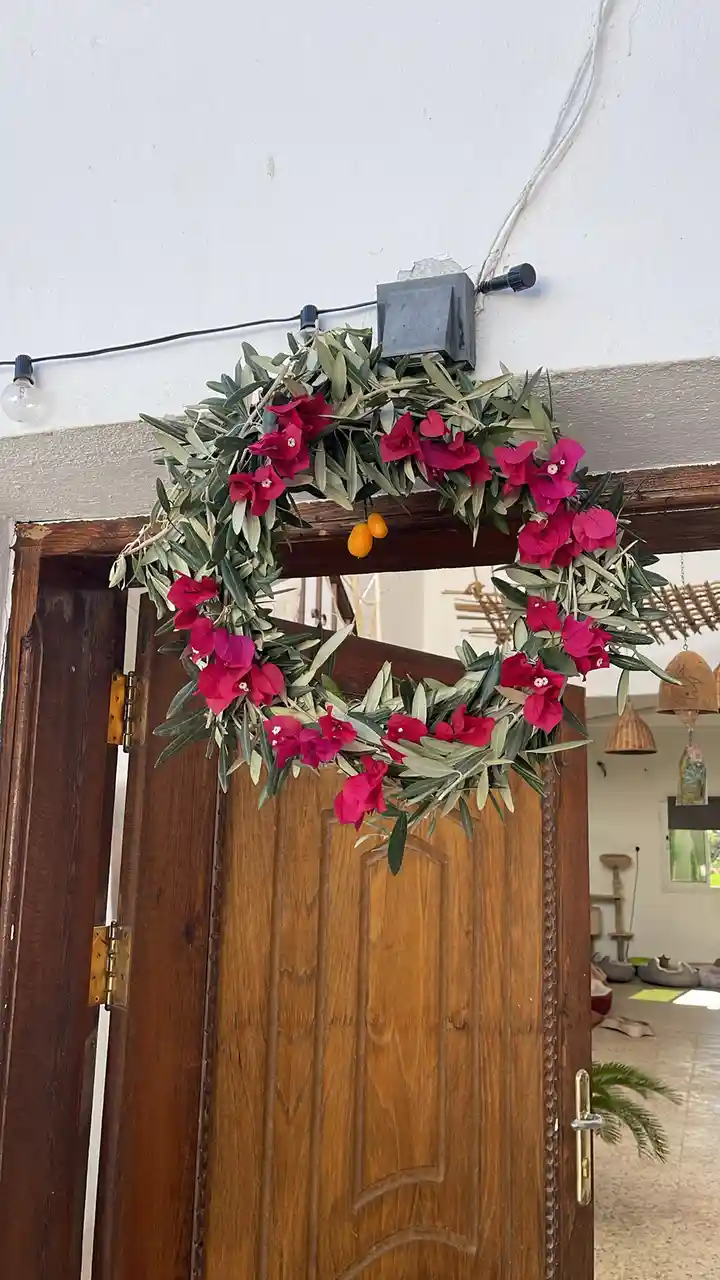 A wooden door with a decorative wreath made of green leaves and bright pink flowers hanging above it. The wreath also…