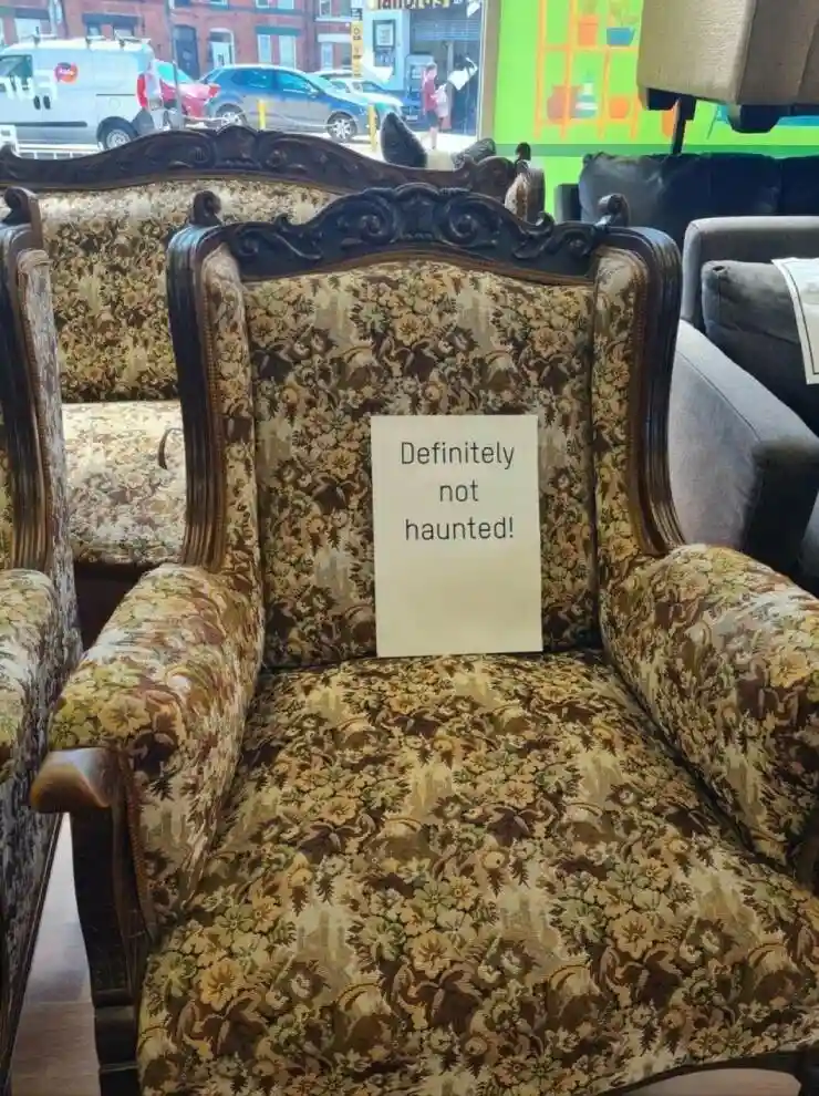 An ornate, floral-patterned armchair in a furniture store has a white paper sign taped to its backrest that reads…