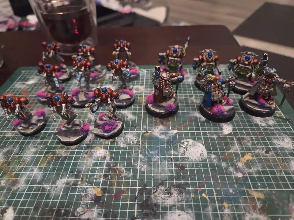 A collection of painted miniature sci-fi robot figures, mostly in red and blue armor, stand on rocky bases with purple…