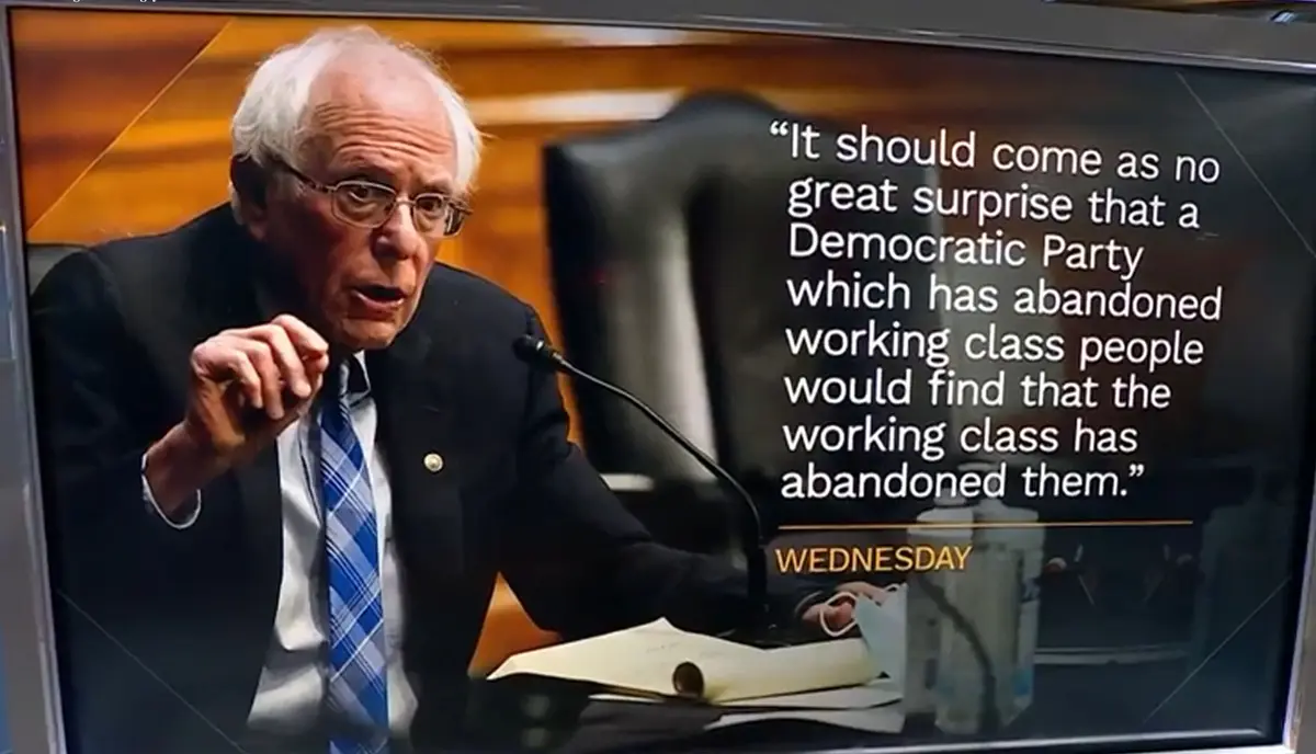 A television screen shows Bernie Sanders speaking at a podium, wearing a suit and blue plaid tie, gesturing with his right…