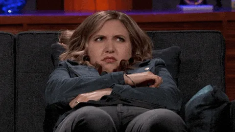 A woman sits on a couch with her arms crossed, showing a progression of facial expressions from skeptical disbelief to…