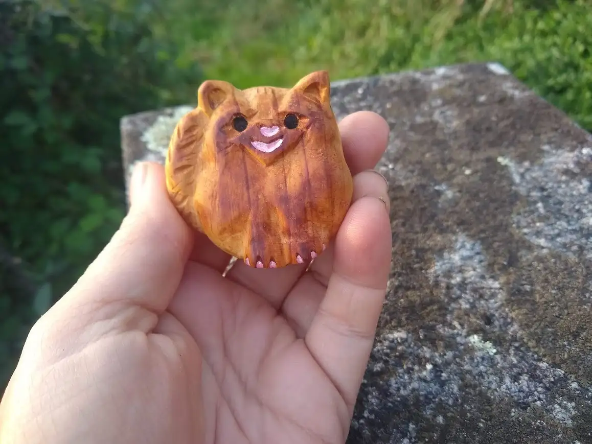 A hand holds a small, carved wooden figurine shaped like a cat with painted black eyes, a pink smiling mouth, and…