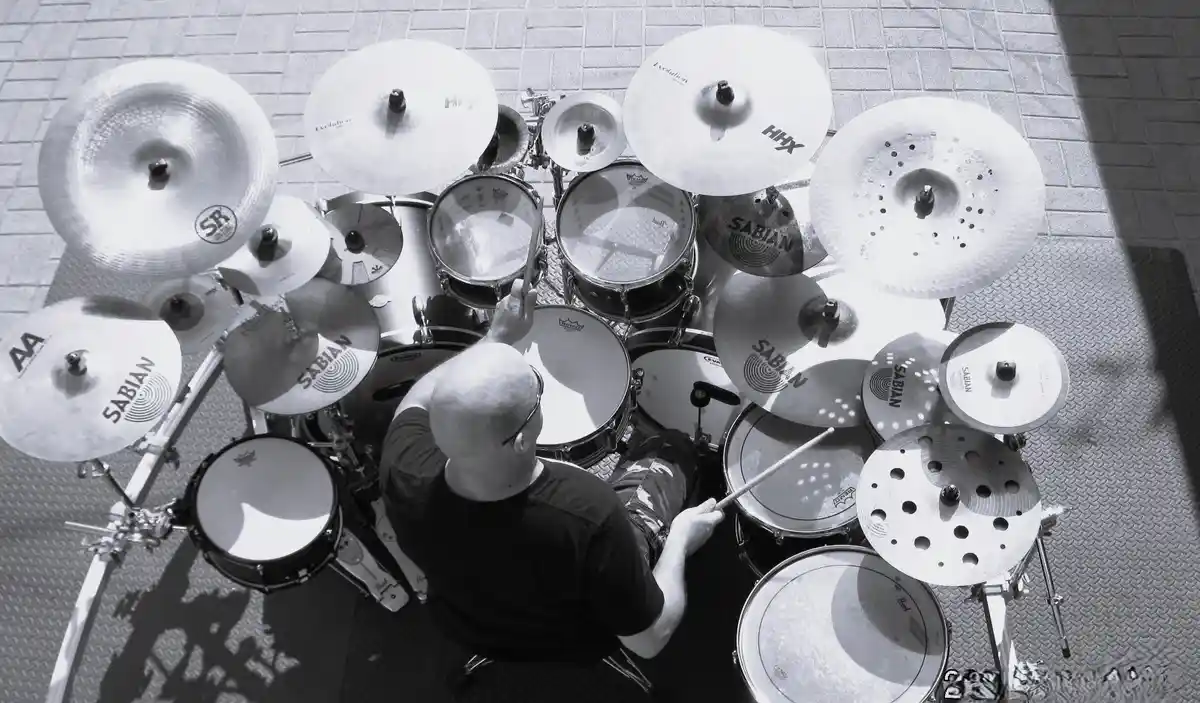 A black-and-white overhead photo shows a bald man in a black t-shirt sitting behind an extremely large, elaborate drum set…