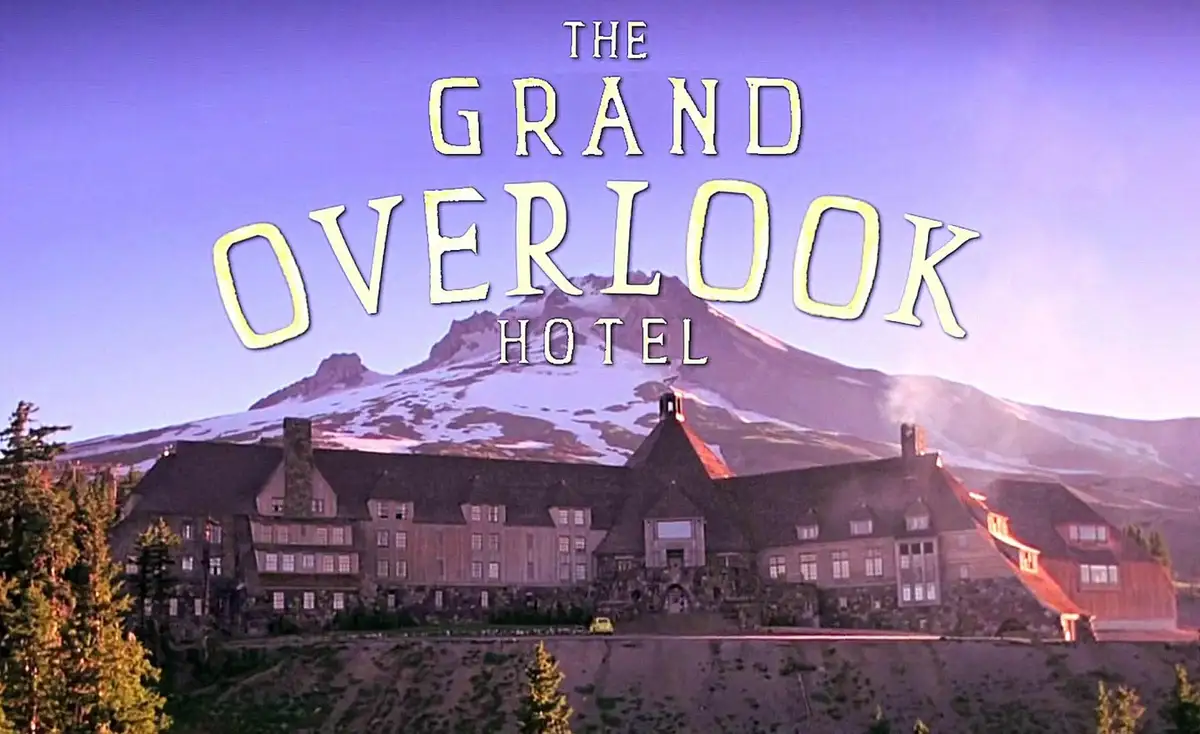 A scenic image of a large, rustic hotel perched on a cliff overlooking a snow-capped mountain range under a purple-hued sky…