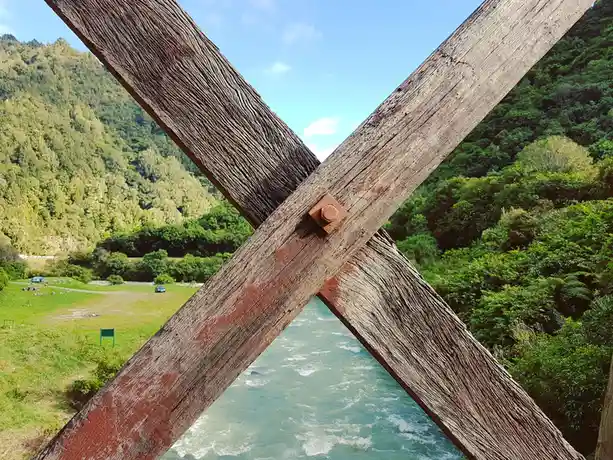A close-up view from a wooden bridge shows two weathered, diagonally crossed beams with a rusty bolt at the intersection…