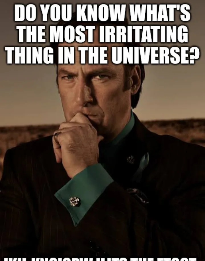 A man in a dark suit with teal shirt cuffs and a ring on his finger, looking thoughtful with his hand on his chin against a…
