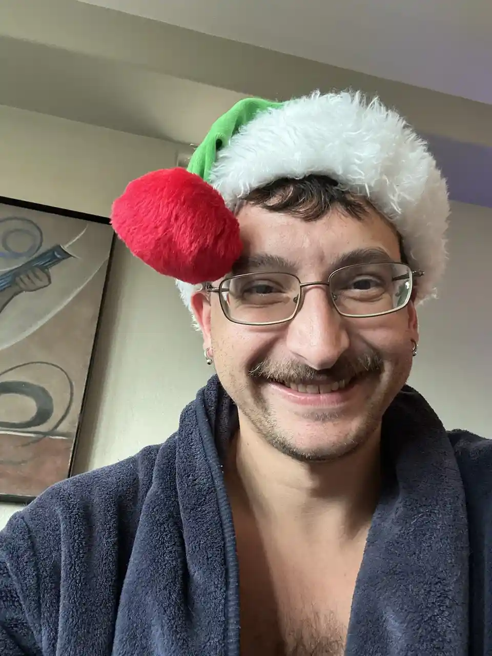 A man wearing glasses and a dark blue robe smiles at the camera while wearing a green and white Santa hat with a red pom-pom…