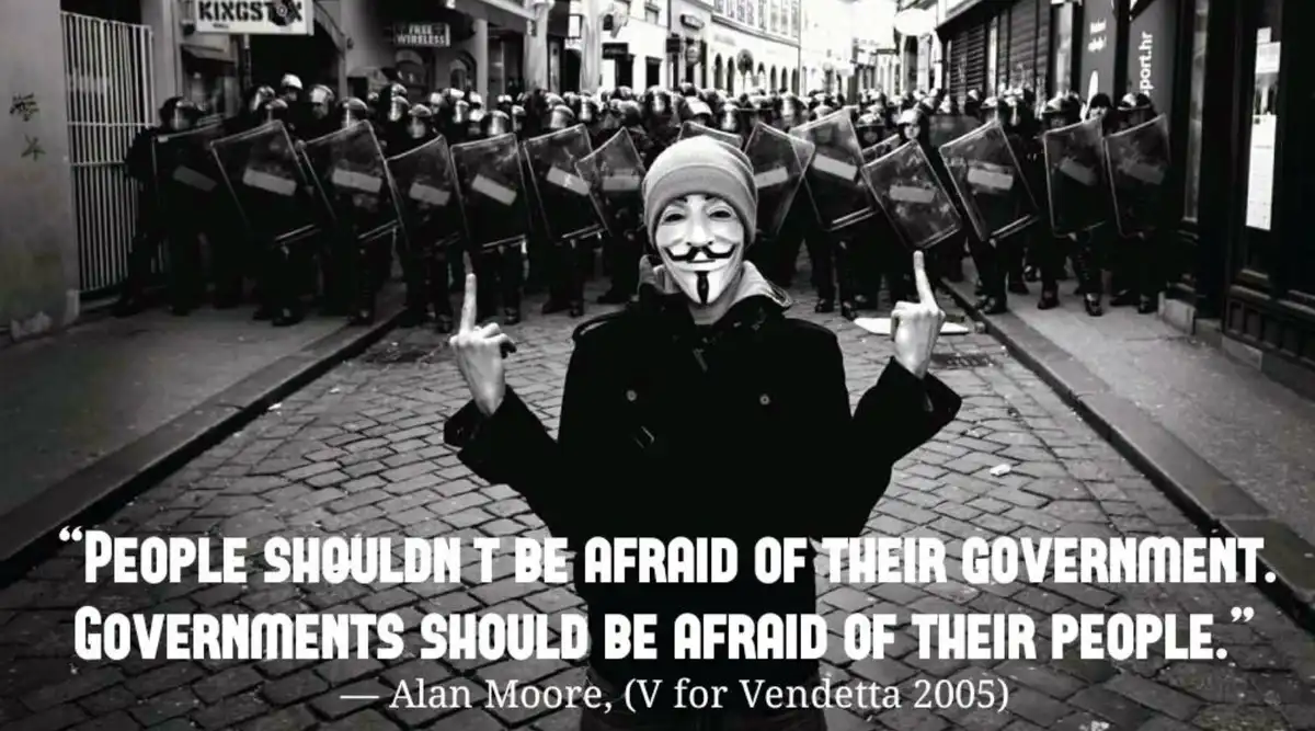 A black-and-white photo shows a person wearing a Guy Fawkes mask and a beanie, standing defiantly in the middle of a…