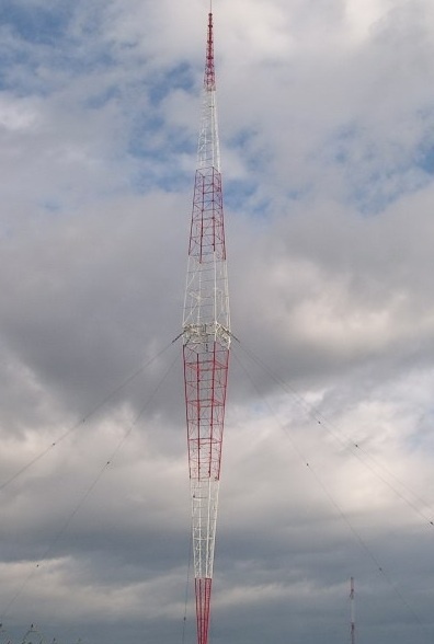 A tall red and white lattice radio tower stands against a cloudy sky, with guy wires extending outward. The image has…