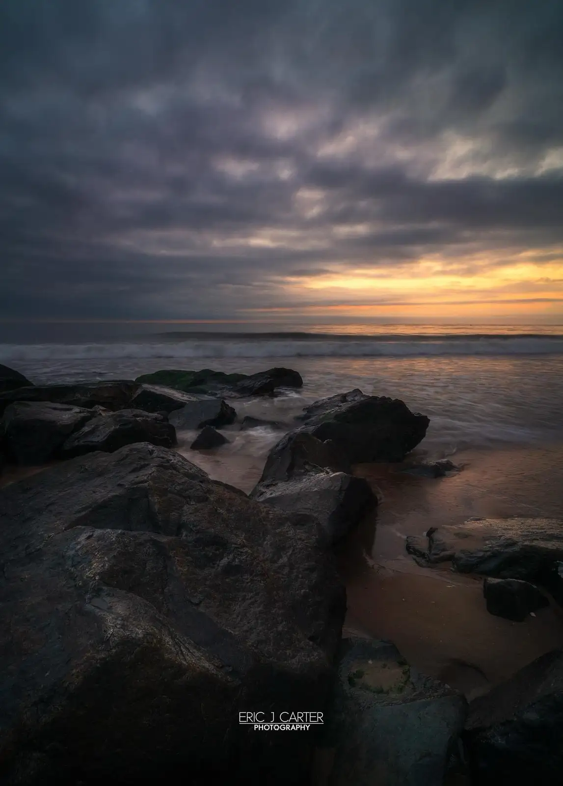 A dramatic long-exposure photograph of a rocky beach at sunset, with dark, textured rocks in the foreground, gentle waves…