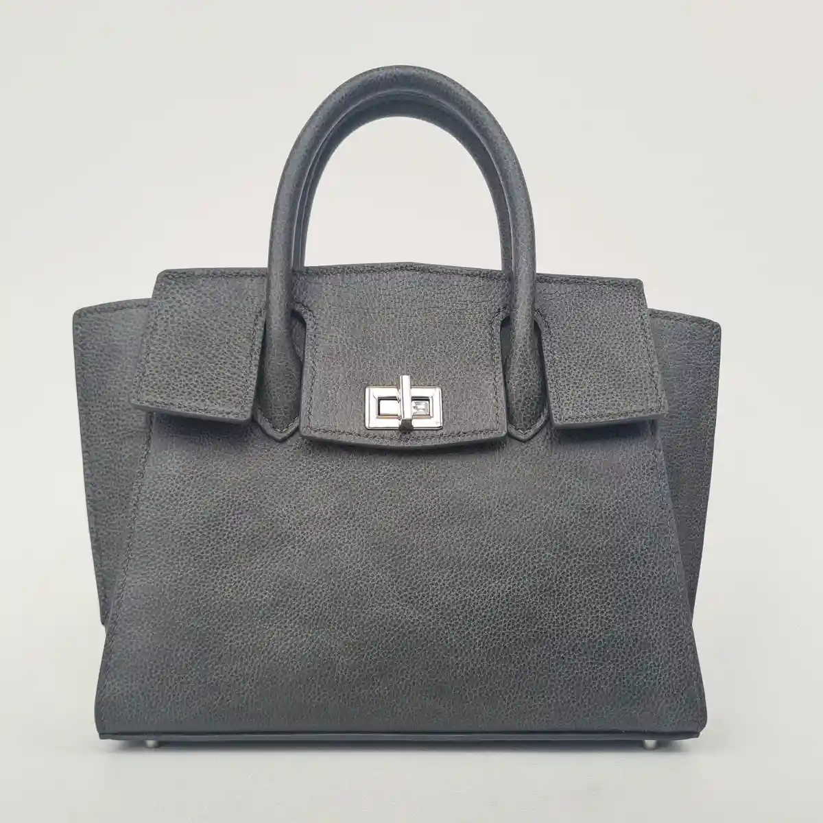 A grey, textured leather handbag with silver hardware and top handles is shown against a plain white background. The…