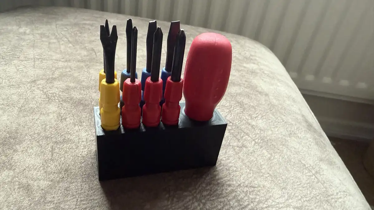 A close-up photo shows a set of screwdrivers with colorful plastic handles (yellow, red, blue) standing upright in a black…