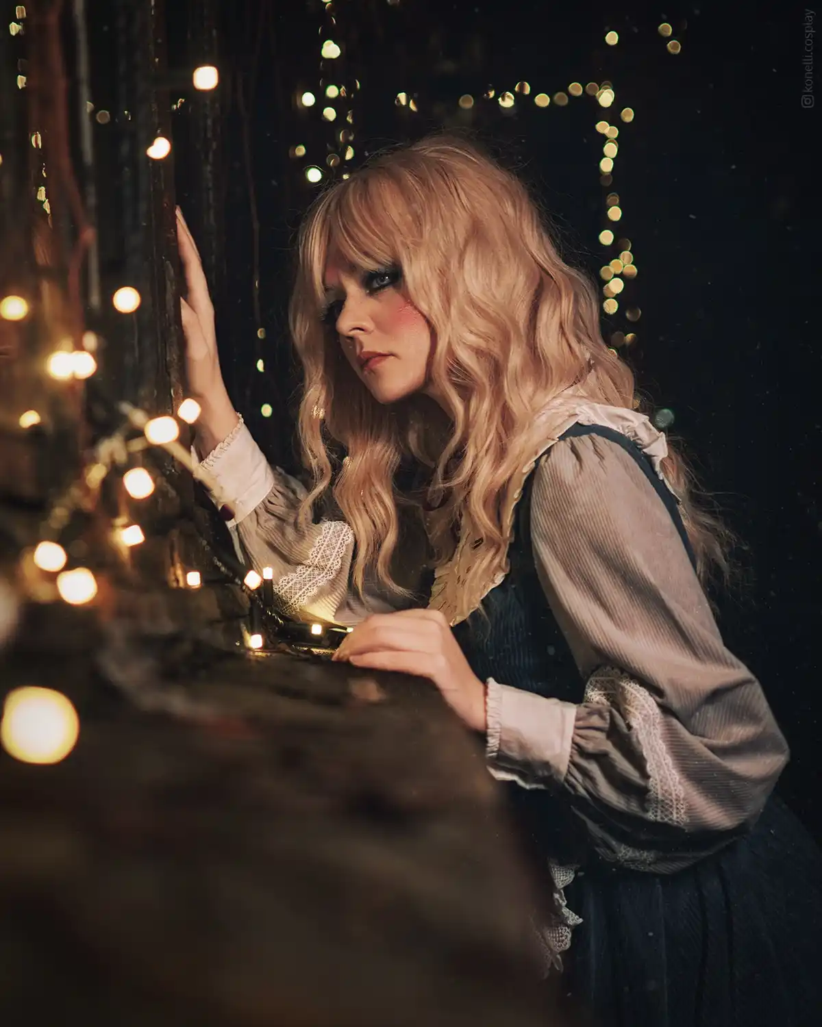 A person with long blonde hair and dramatic makeup, wearing a vintage-style blouse and dark overalls, leans against a wooden…