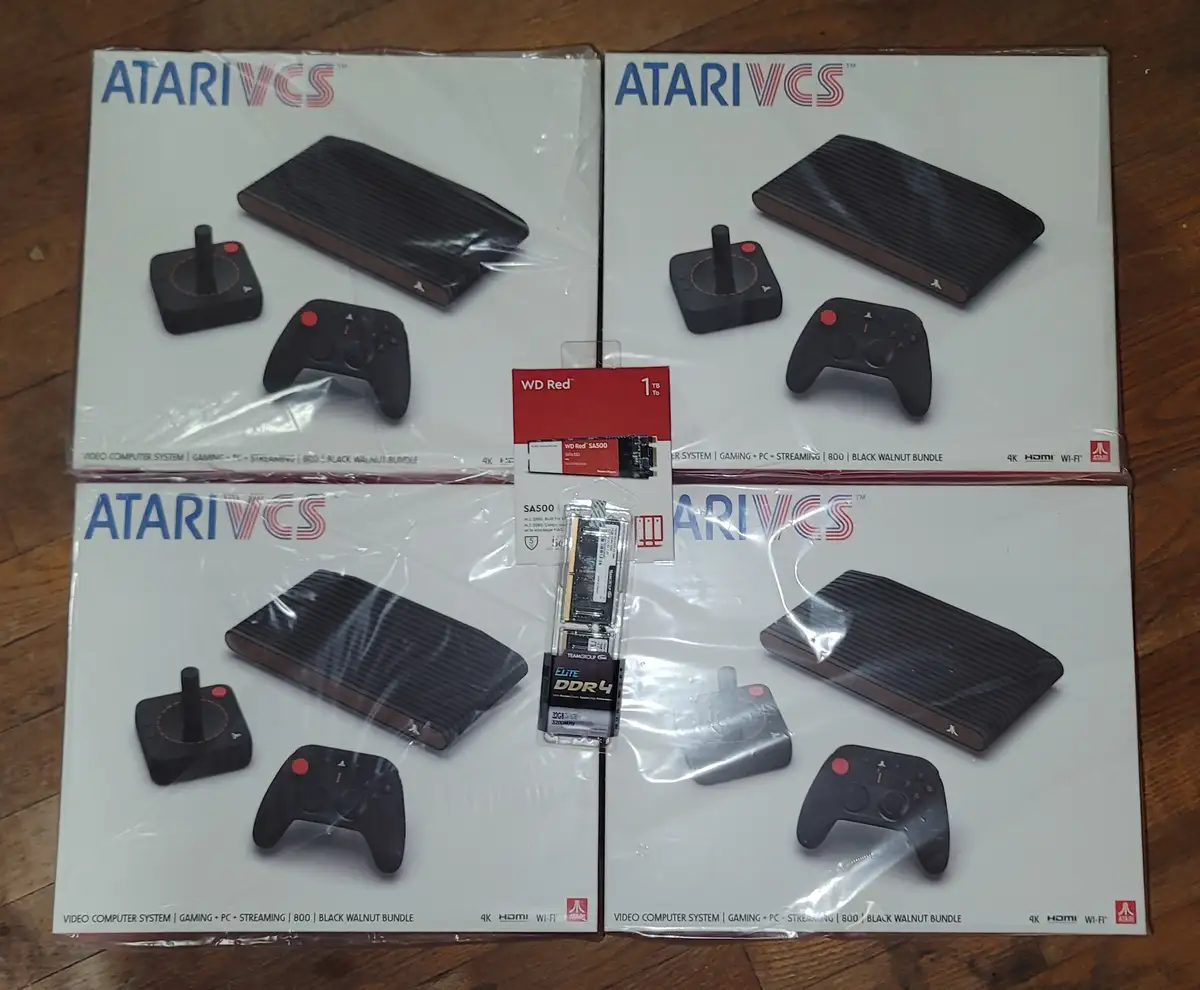 Four identical Atari VCS console boxes are arranged in a two-by-two grid on a wooden surface, with a WD Red 1TB SSD and a…