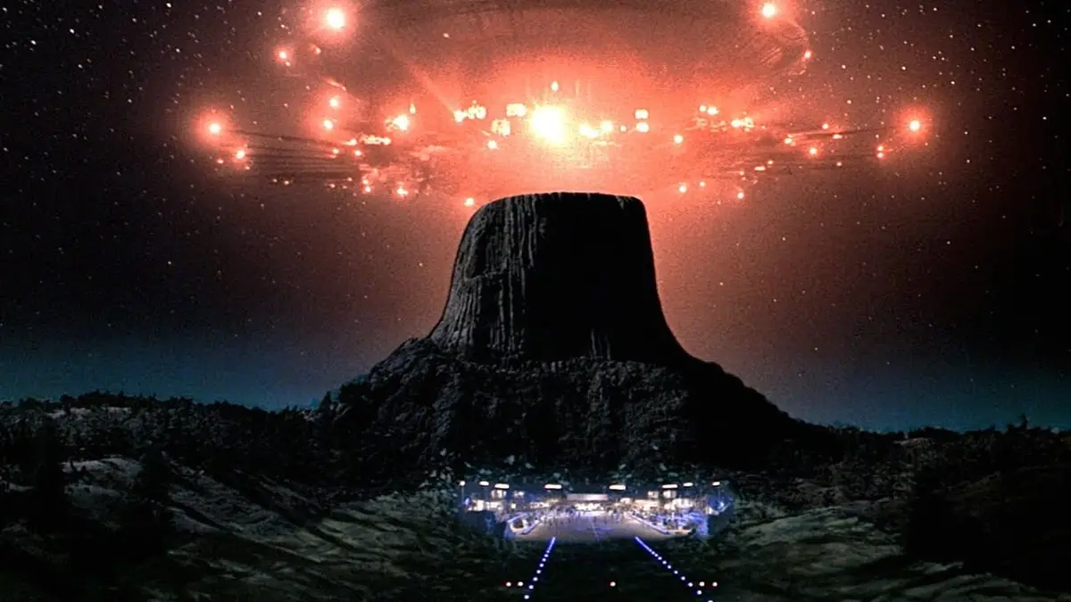 A night scene featuring Devils Tower under a starry sky, with a glowing red UFO hovering above it. Below, a brightly lit…