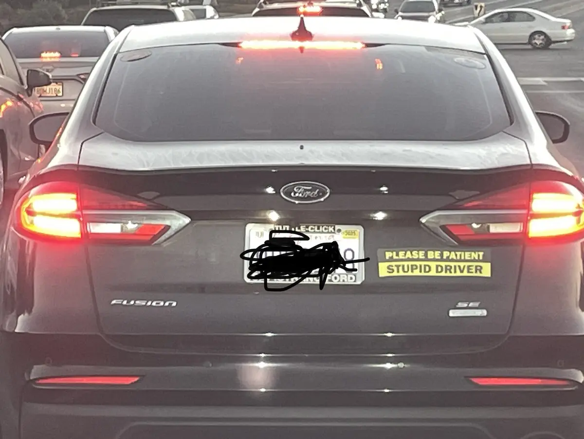 The rear of a dark gray Ford Fusion is shown in traffic with its brake lights on. The California license plate is obscured…