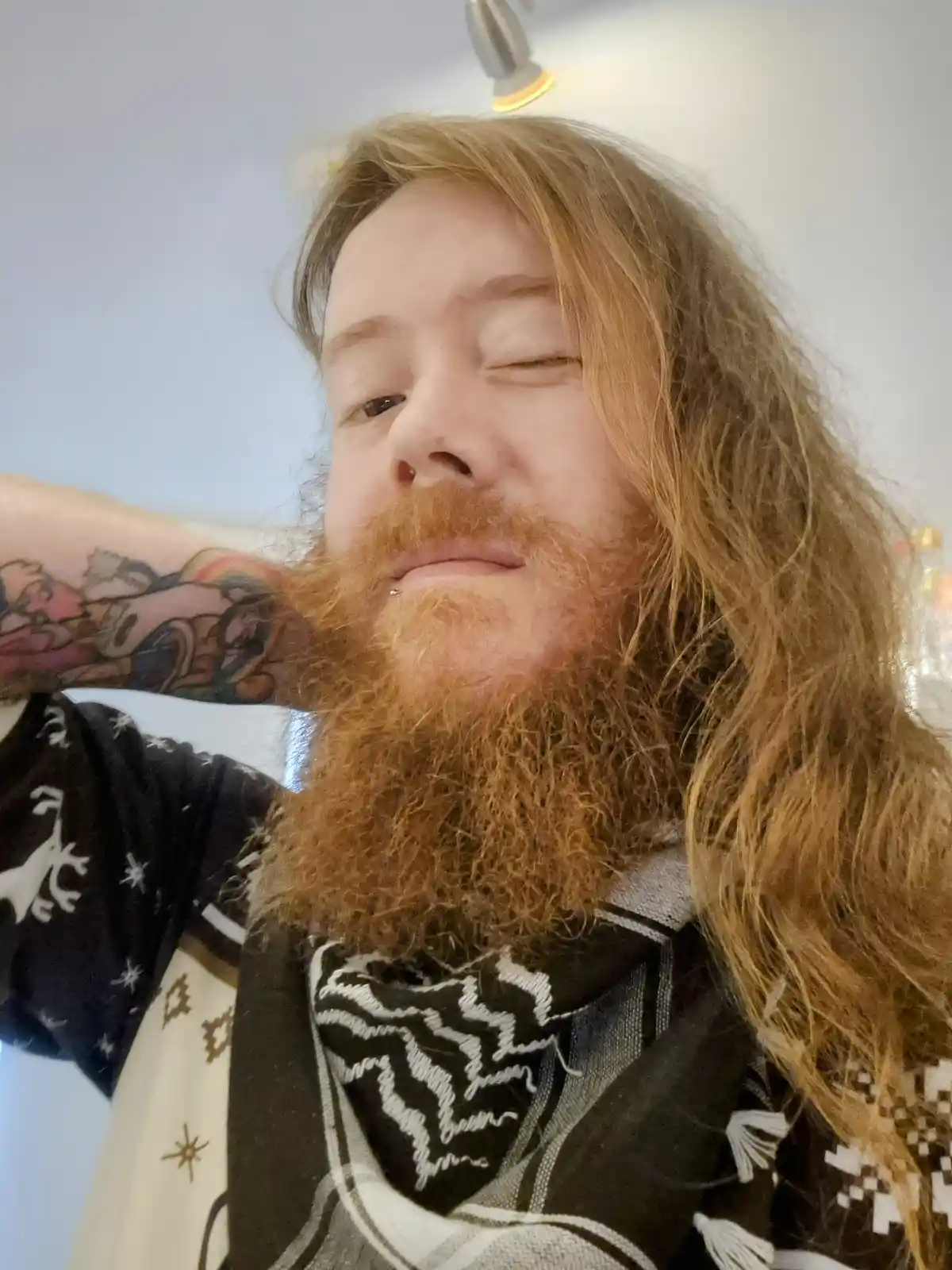 A person with long, reddish-brown hair and a thick, full beard is shown in a selfie, winking one eye while the other is…