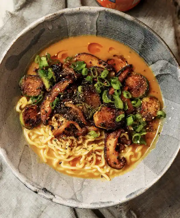 a top-down photo of a rustic ceramic bowl filled with ramen noodles in a rich broth, topped with roasted brussels sprouts…
