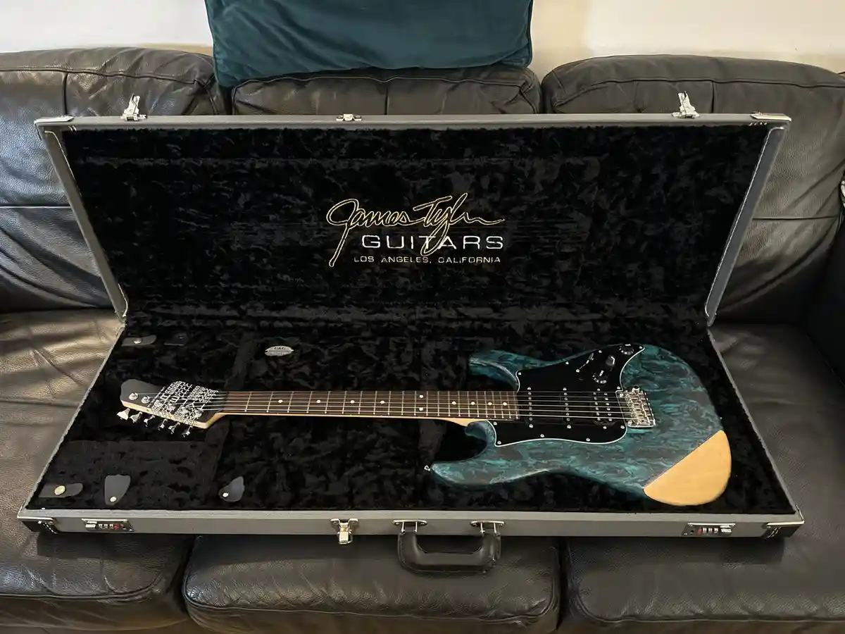 A teal and black electric guitar with a light wood pickguard rests inside an open, black velvet-lined hard case branded…