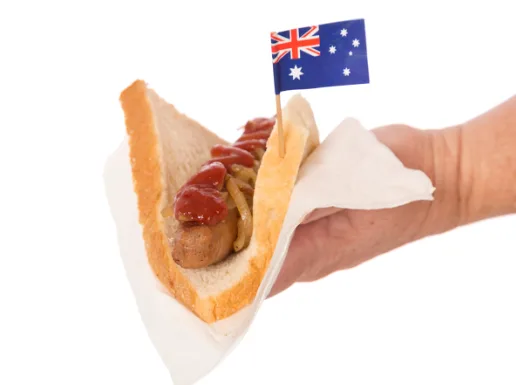 A hand holds a hot dog in a bun with ketchup and onions, topped with a small Australian flag on a toothpick. The image is…