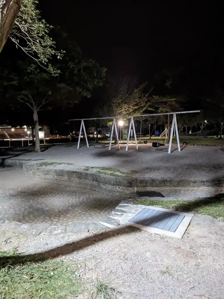A nighttime photo of an empty playground with swings, illuminated by a single light, showing a gravel path and a drain grate…