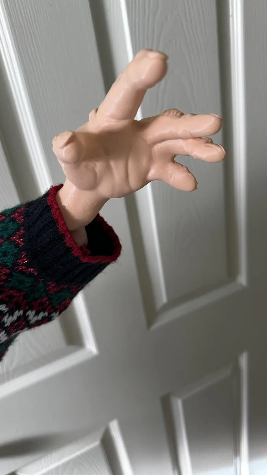 A person wearing a festive red, green, and black patterned sweater has their hand stuck in a white paneled door, with the…