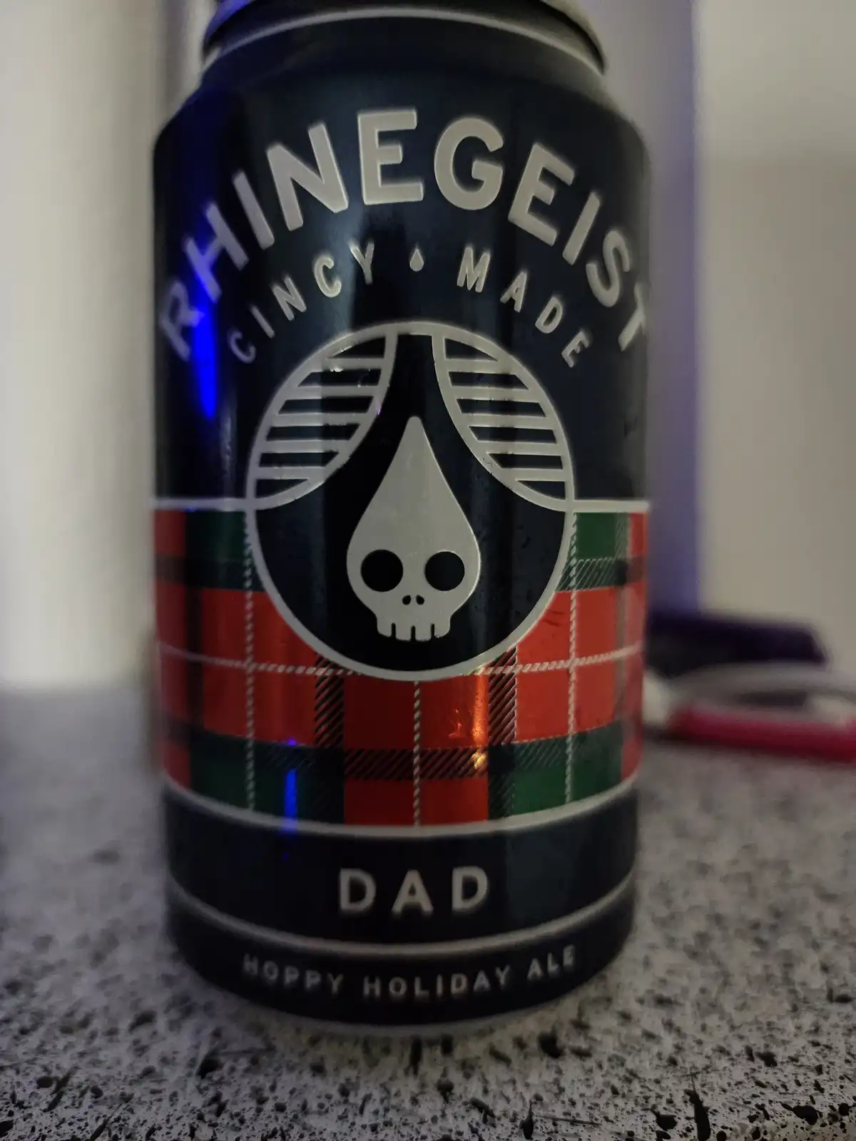 A close-up photo of a black beer can with a skull logo inside a circle, surrounded by a red, green, and black plaid pattern…