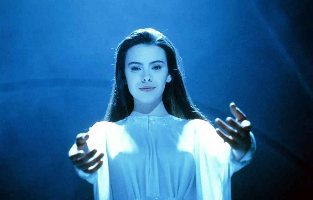 a woman with long dark hair wearing a white robe stands with her hands open, bathed in blue light. The overlaid text reads…