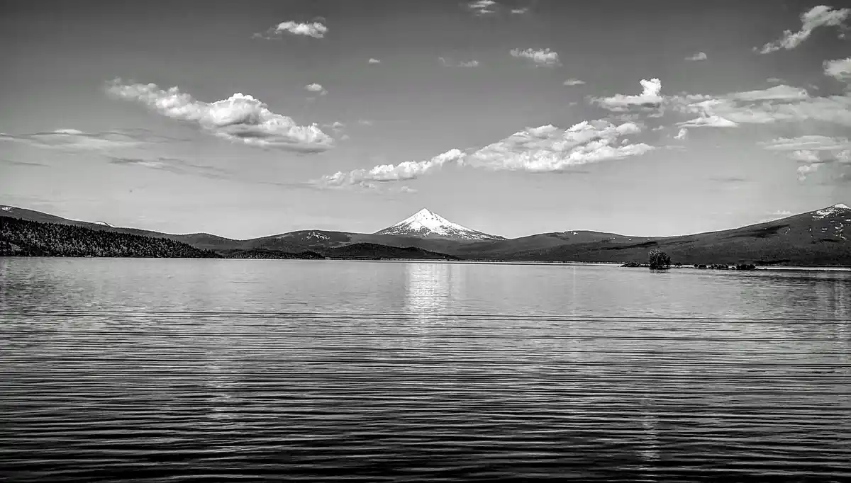 A black-and-white photograph shows a calm lake with ripples, surrounded by forested hills and a prominent snow-capped…