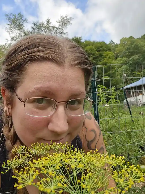 A person with glasses and a tattooed arm holds a large bunch of yellow dill flowers in front of their face, obscuring their…