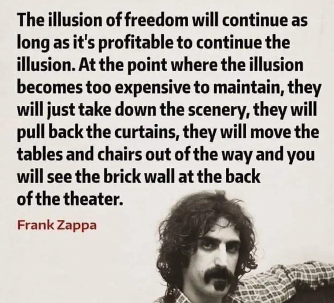 A black-and-white photo of Frank Zappa with long curly hair and a mustache, wearing a plaid shirt, looking toward the…