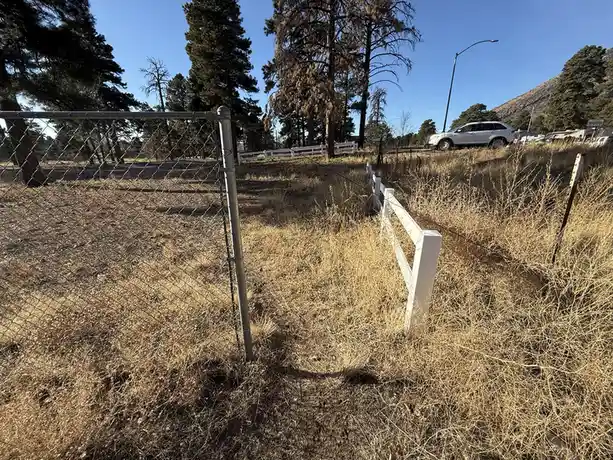An outdoor photo shows a chain-link fence and a white picket fence in a dry, grassy field with trees and a white SUV in the…