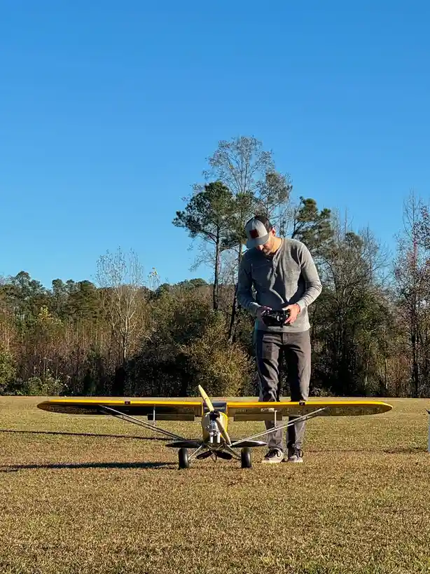 A man in a grey long-sleeve shirt and baseball cap stands in a grassy field, looking down at a remote control in his hands…