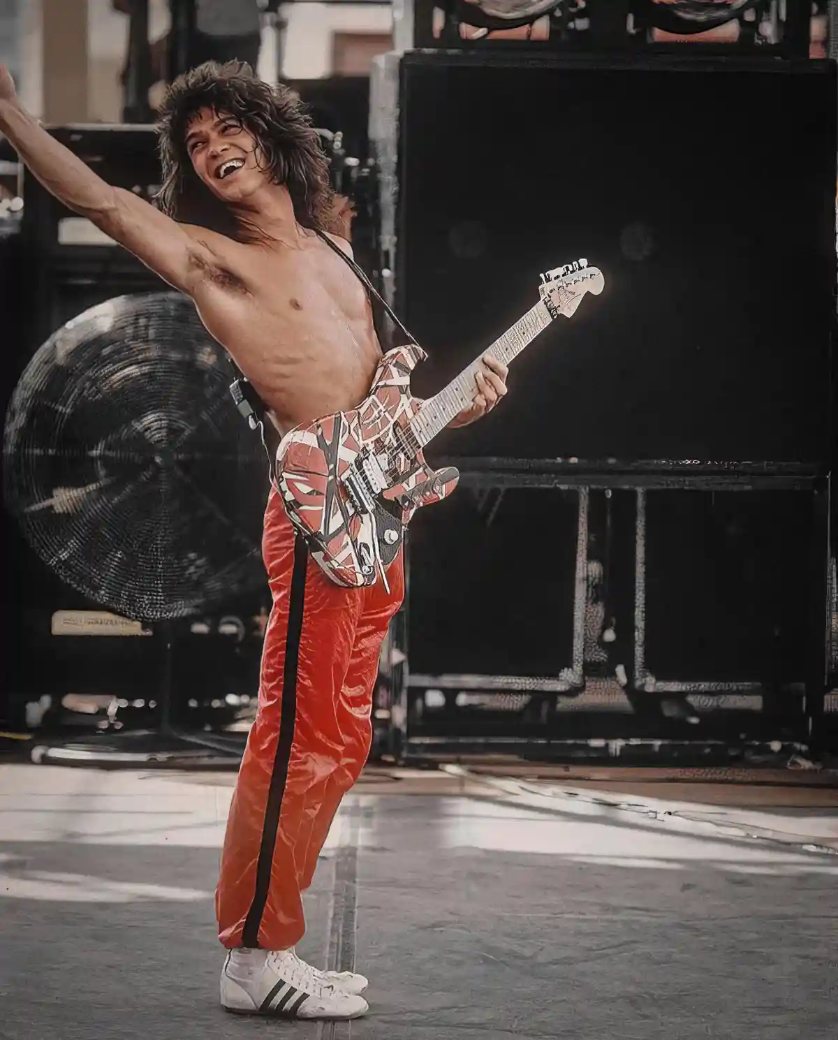 A shirtless man with long hair, wearing red pants and white sneakers, joyfully plays a guitar with a distinctive red, white…