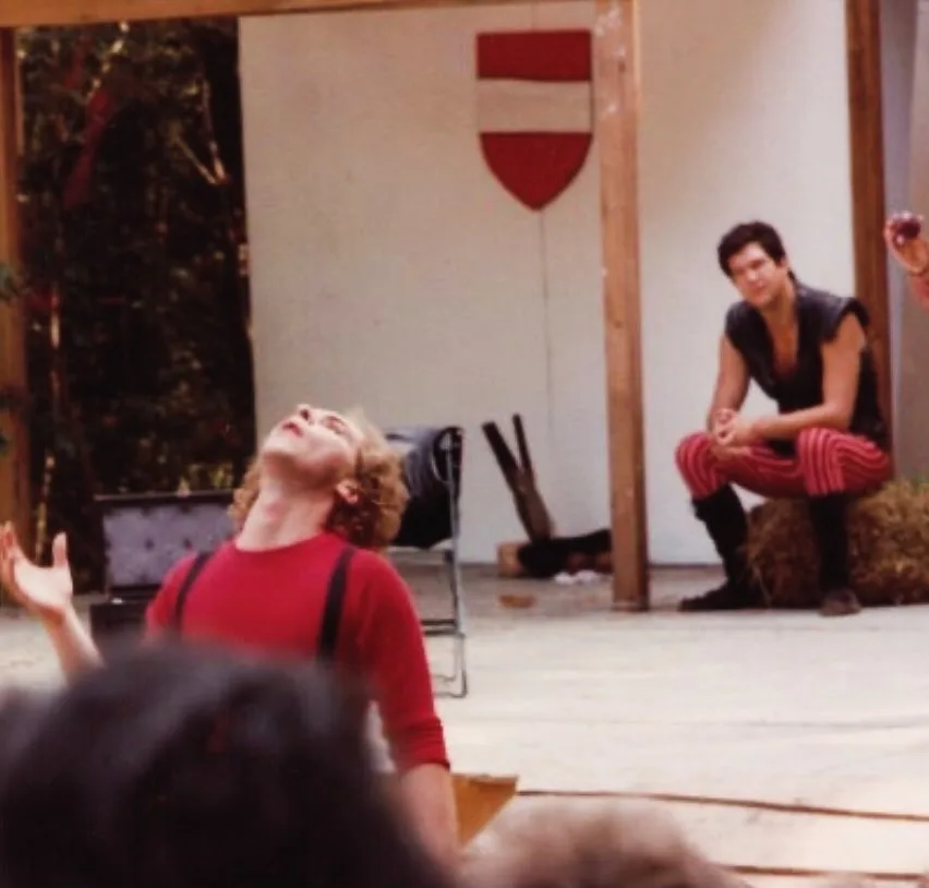 A man in a red shirt and suspenders is dramatically tilting his head back with his arms outstretched, as if in despair or…