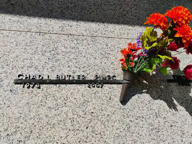 A granite memorial plaque for Chad L. Butler, PIMPC, with birth and death years 1973 and 2007, has a bouquet of artificial…