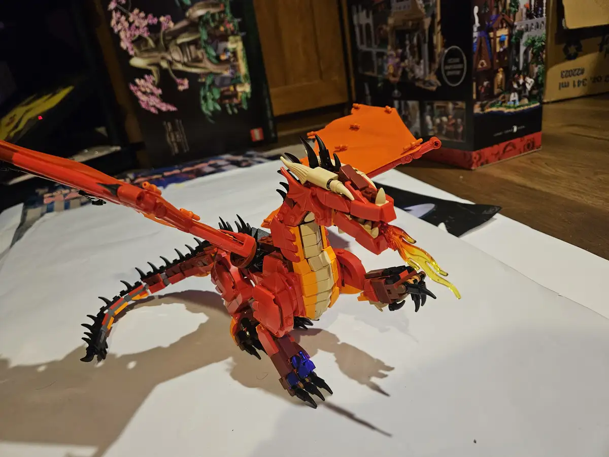 A red and orange LEGO dragon model with black spikes, wings, and flame accents is posed on a white surface. In the…