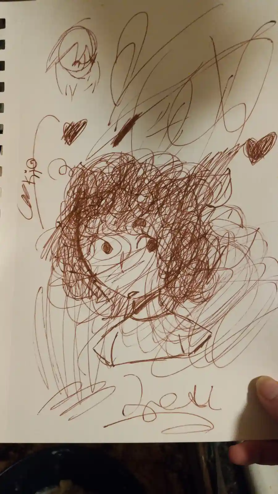 A messy brown ink drawing on spiral notebook paper shows a crudely sketched anime-style character with scribbled hair and…