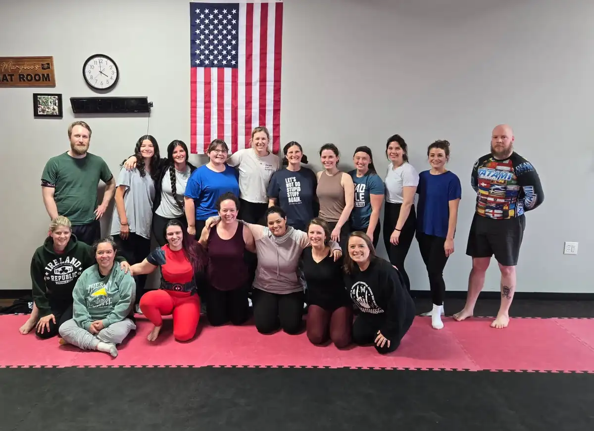 A group of people, mostly women, pose for a photo in a gym with an American flag on the wall. One woman in the center wears…