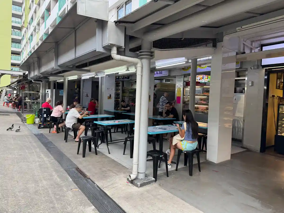 An outdoor hawker center scene with people sitting at blue tables eating, a woman in a blue top and light shorts sits alone…