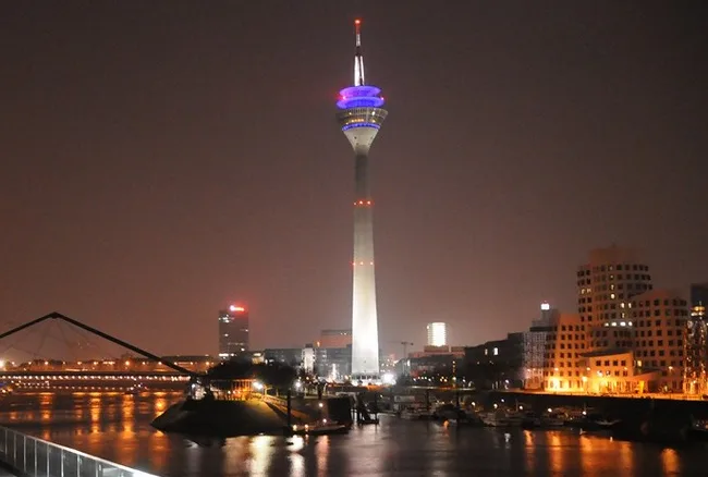 A nighttime photo of the Köln TV Tower (Kölnturm) in Germany, illuminated with blue and purple lights at its observation…