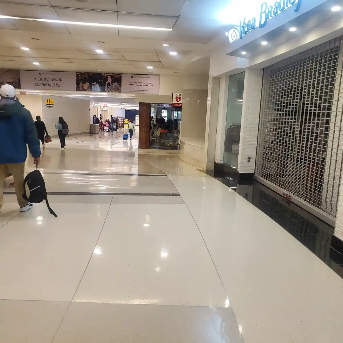 An airport terminal hallway with shiny white floors, travelers walking, and a closed Vera Bradley store with a metal…