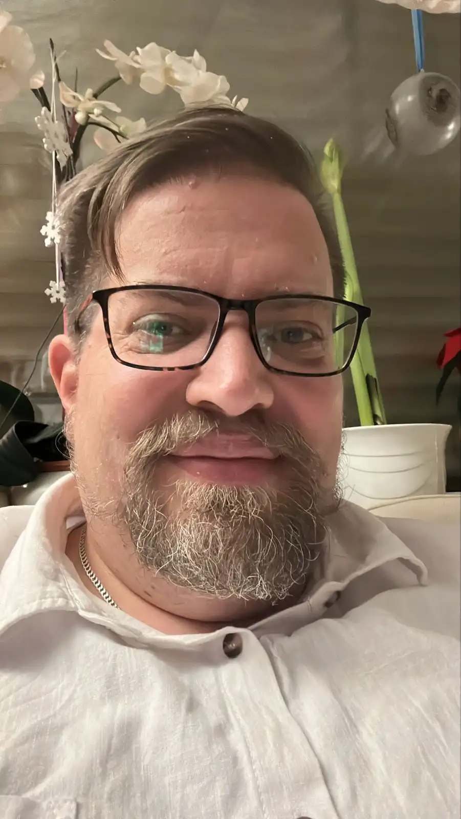A man with glasses and a graying beard, wearing a white shirt and a silver chain, looks at the camera with a slightly…