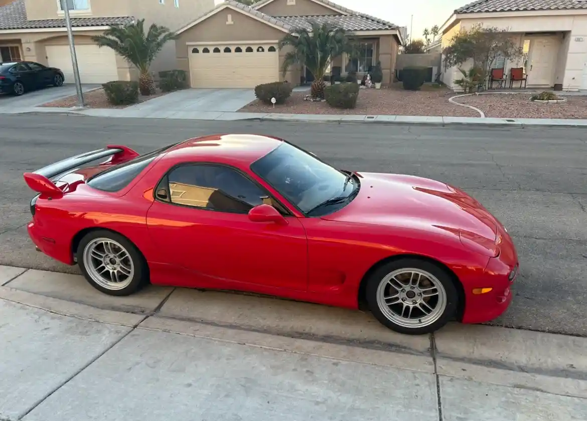 A red 1990s Toyota Supra is parked on a residential street curb, with a suburban neighborhood featuring houses and palm…