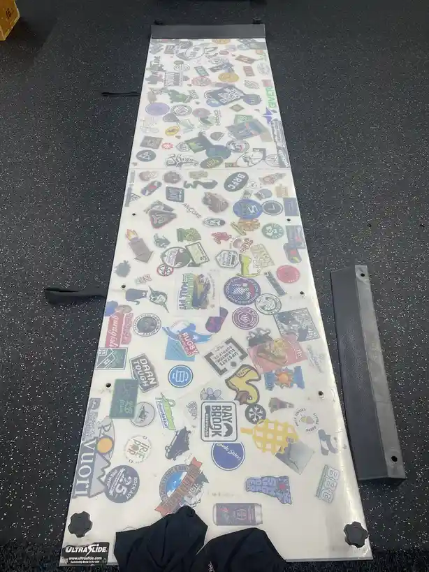 A snowboard covered in hundreds of colorful, overlapping stickers lies on a speckled black floor, with its black plastic…