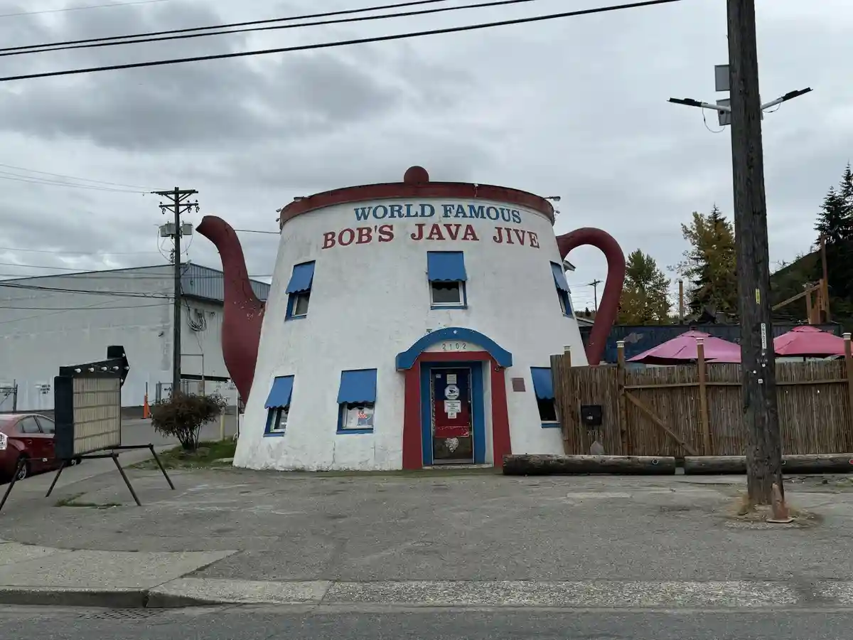 A large, white building shaped like a giant teapot with red spout and handle, featuring blue window awnings and a blue…