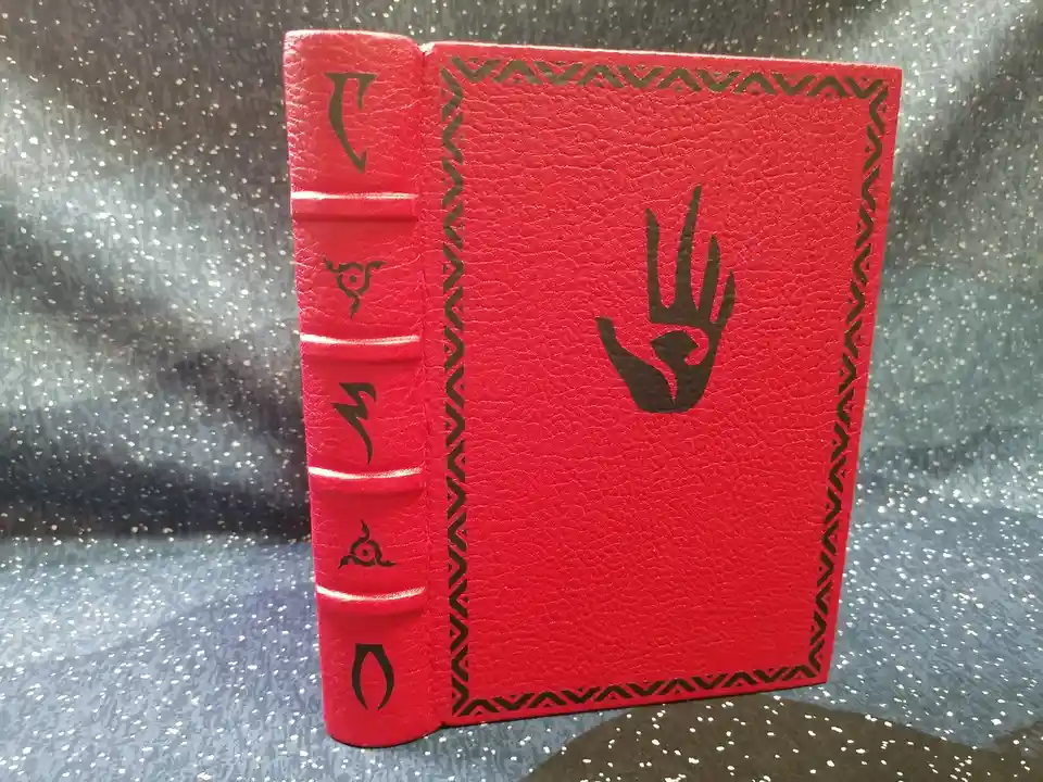 A red leather-bound book with black symbols on its spine and a hand symbol on the cover, standing upright on a speckled…