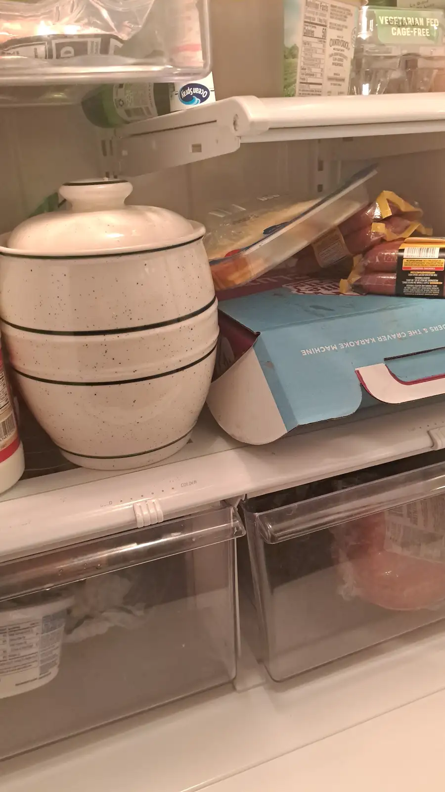 A close-up shot of a refrigerator shelf holding a white speckled ceramic crock with a lid, a blue box labeled 'THE CRAVER…