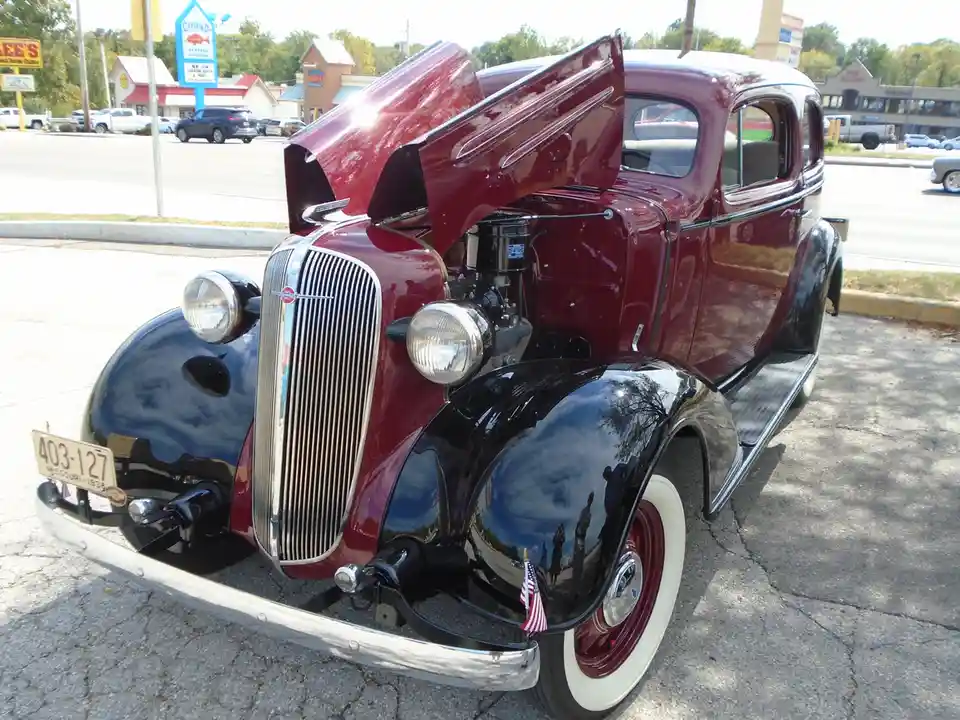 A shiny, two-tone maroon and black 1938 Chevrolet sedan with its hood open, parked in a modern parking lot. The car has a…