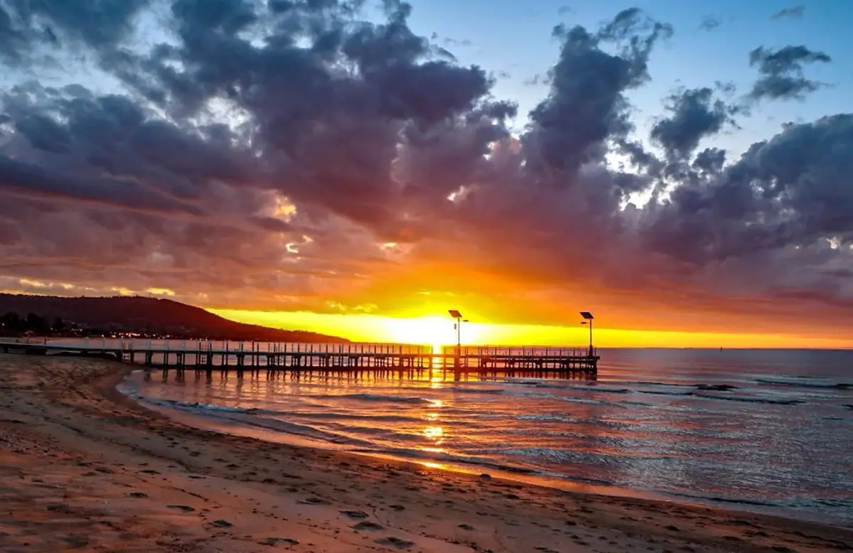 A serene sunset over a beach with a long pier extending into the water, the sky painted with vibrant orange, yellow, and…